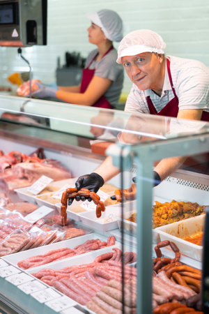 Male seller leans over to refrigerator window and takes out some raw barbecue sausages for buyerの写真素材