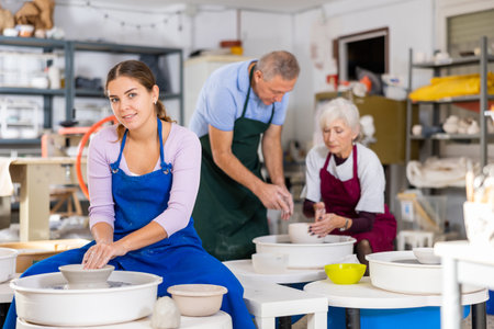 Female positive artisan in ceramics workshop with pottery wheel and various clay vesselsの写真素材