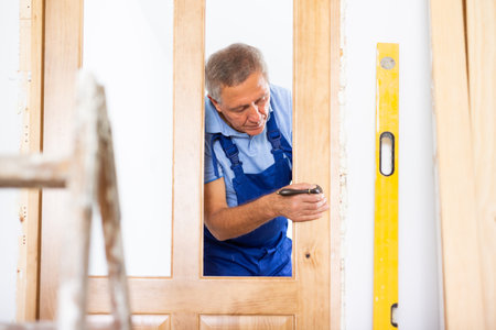 Professional middle-aged repairman in uniform installing a door handle on a door during renovation, indoorsの写真素材