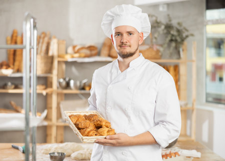 Baker in white uniform stands behind counter in small private bakery offering hot croissantsの写真素材