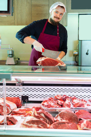 Young bearded butcher slicing fresh beef for customer at counterの写真素材