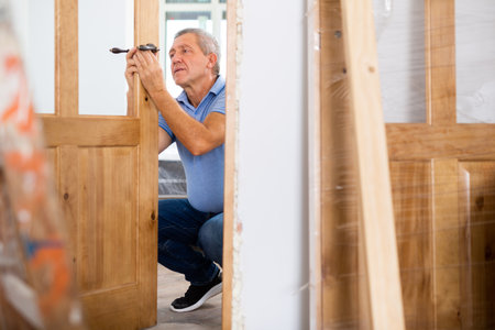 Professional middle-aged repairman installing a door handle on a door during renovation, indoorsの写真素材