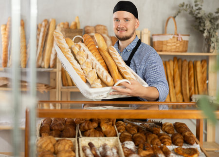 Male seller stands at bakery counter with basket filled by fresh baguettes.の写真素材