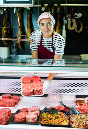 Female butcher shop vendor offering burger patties from display caseの写真素材