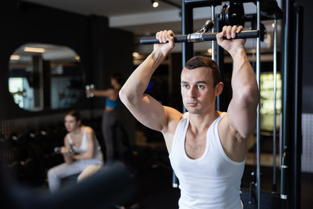 Slender athletic man work out muscles of arms and shoulders on simulator in gym of fitness club