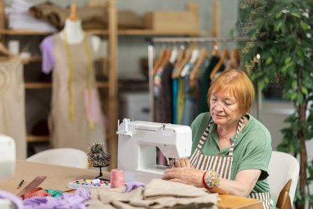 Mature female seamstress is sitting near sewing machine and create new dress in workshopの写真素材