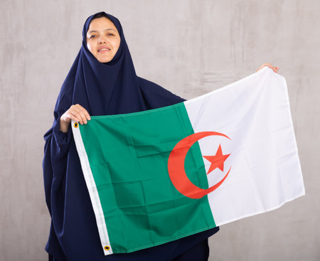 Young Muslim woman in blue hijab holds unfurled flag of Algeria.の写真素材