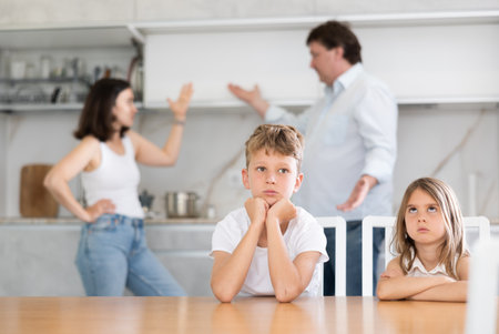 Two children sitting at the kitchen table sadly while their parents quarrellingの写真素材