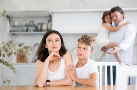 Offended mother sitting at the kitchen table being hugged by sonの写真素材