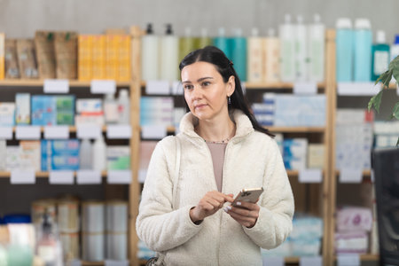 Woman is standing in a pharmacy and holding a mobile phone in her handsの写真素材