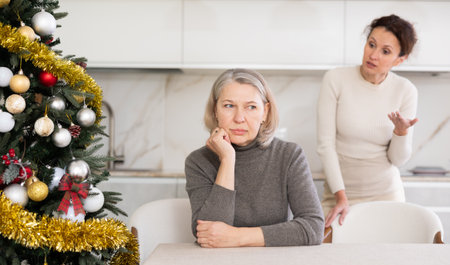 Old woman sitting at table while middle-aged female family member scolding her in the kitchen with Xmas-treeの写真素材