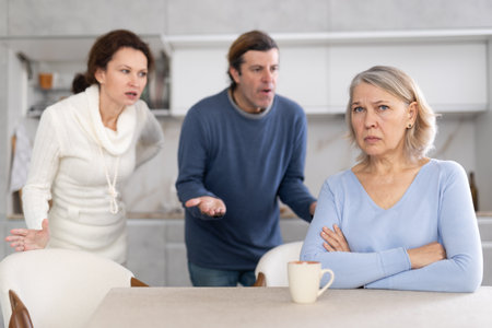 Woman and man swear at old mother who puts up with it while sitting at kitchen tableの写真素材
