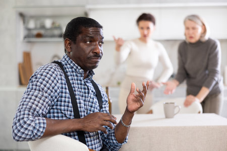 Middle-aged man sitting at the kitchen table with his back to women quarreling to himの写真素材