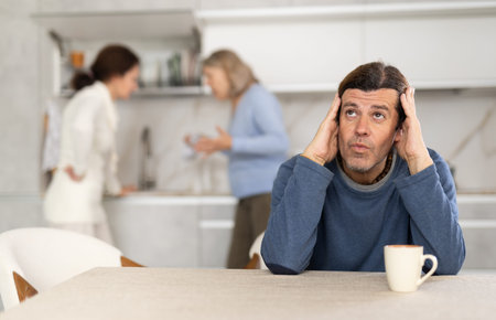 Frustrated man sitting in kitchen during quarrel between his wife and elderly mother.