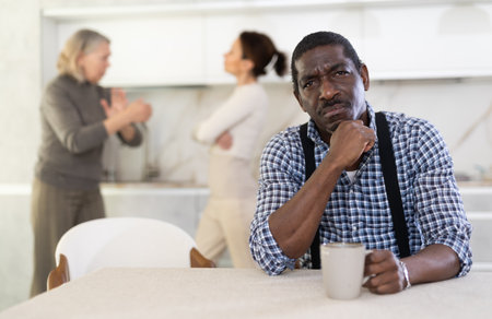 Middle-aged man sitting at the kitchen table with his back to women quarreling togetherの写真素材