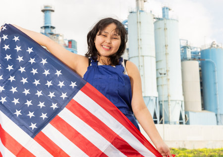 Happy asian girl in work clothes with flag of usa standing in front of industrial sceneryの写真素材