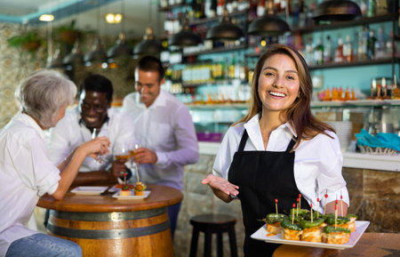 Cheerful smiling female waiter holding served tray meeting visitors at barの写真素材