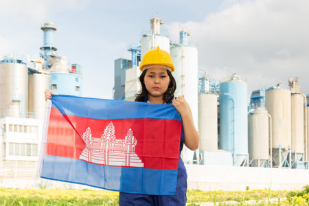 Sad young woman in helmet holding flag of Cambodia against background of factoryの写真素材