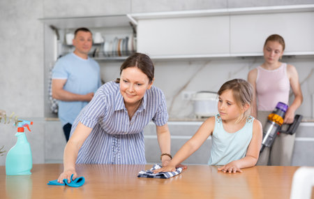 Cheerful woman wiping table in kitchen with preteen daughterの写真素材