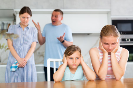 Frightened and frowning children sit at kitchen table during mom and dad quarrel.の写真素材