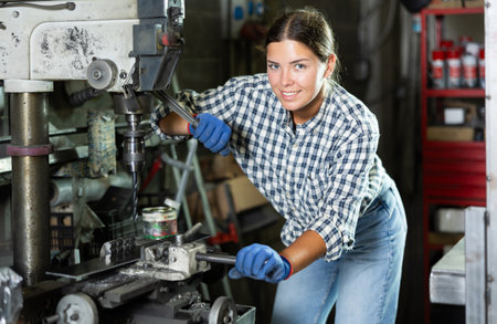 Young woman using drill press to hole metal part in workshopの写真素材