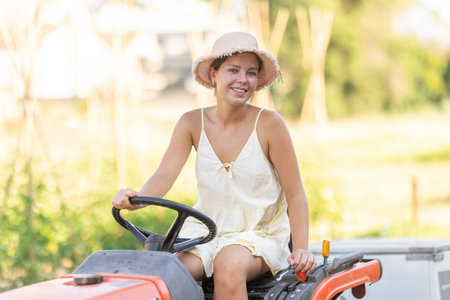 Young woman sits on minitractor and drives rural vehicle, performs loading work on farmの写真素材