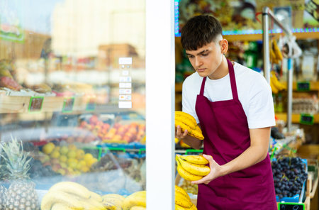Grocery store employee in an apron puts fresh bananas on shelvesの写真素材