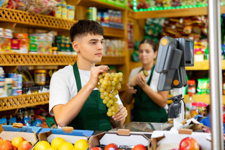 Young male supermarket worker weighing on the scales green grapes in grocery shop