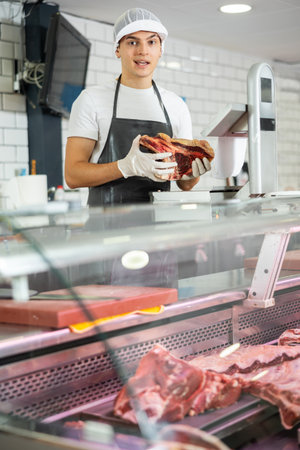 Positive young salesman demonstrating piece of meat in butcher shopの写真素材