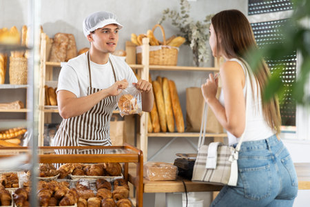 Young man seller puts croissants in bag to woman buyer