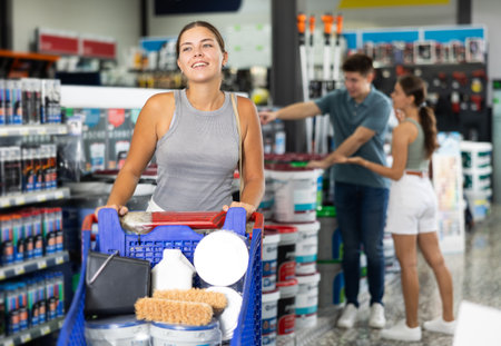 Girl with trolley is standing in sales area of paintwork material department of supermarket recalls position of goods needed for purchase. Male and female buyers in backgroundの写真素材