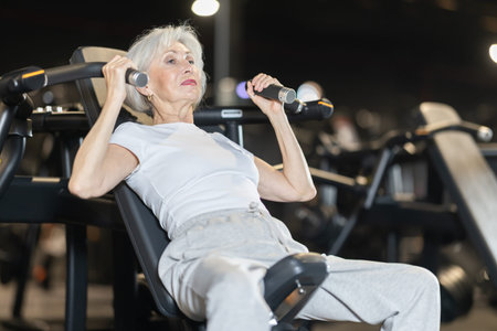 Sporty elderly woman using shoulder press machine at gym