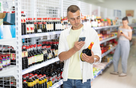 Male buyer examines packaging of product and scan QR code on label soy sauces in supermarketのeditorial素材