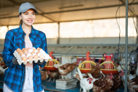 Smiling woman in plaid shirt and cap collecting eggs in chicken farm
