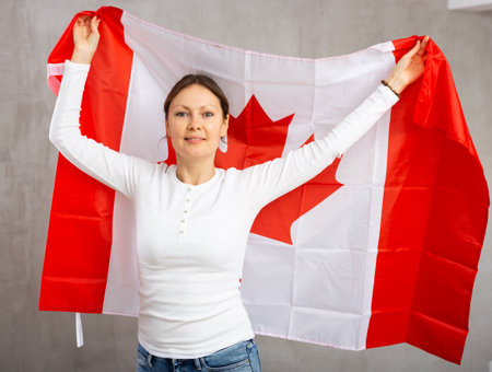 Smiling relaxed woman waving national flag of Canada while looking at camera at gray wall backgroundの写真素材