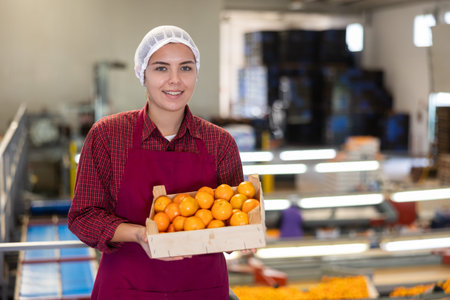 Glad positive female employee in colored uniforms hold a box of fresh ripe tangerines in their hands...