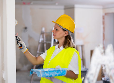 Professional female painter in protective helmet and yellow vest paints the walls with paint roller