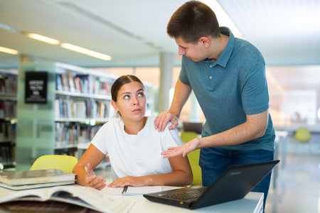 Friendly young woman offering assistance to male friend studying, finding information at library