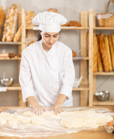 Young female baker kneading dough on table