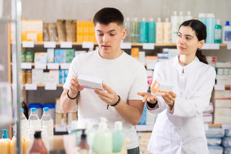 Guy chooses pills in modern pharmacy. Female pharmacist helps with choosing medicineの写真素材