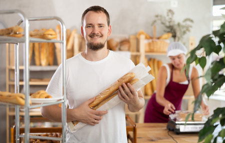 Positive young guy holding baguettes in plastic bag in bakery