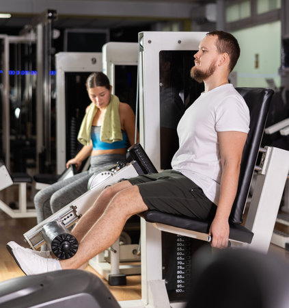 Bearded man in gym performs exercises for leg muscles using simulators and fitness center equipment.