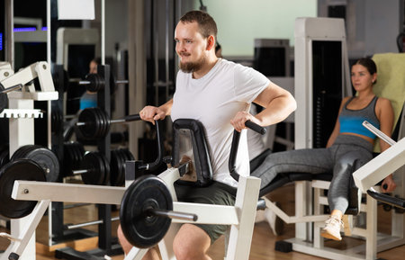 Bearded man in gym performs exercises for arm muscles using simulators and fitness center equipment.