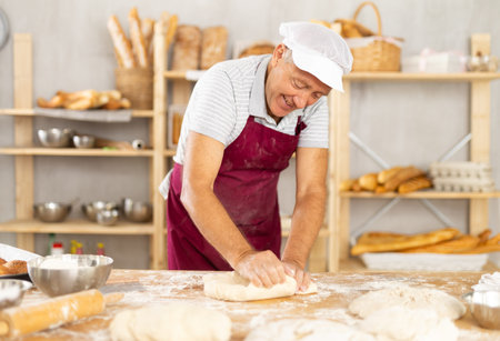 Elderly male baker in uniform prepares raw dough, kneads it with hands