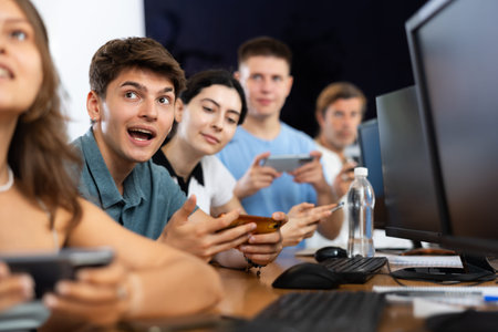 At computer class, young guy student sits with mobile phone and listens to teacher