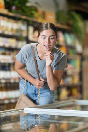 Young woman buyer making choice leaning over shop-fridge in supermarket