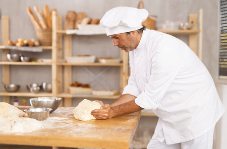 Male baker stands at work bench, kneading and shaping dough to make bread, croissants and baguettes