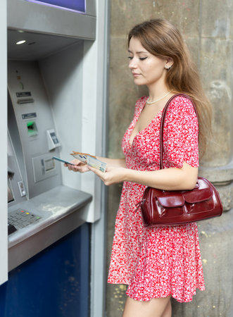 Woman uses ATM, withdraws cash from card. Girl buys travel ticket from machineの写真素材