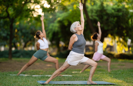 Sporty energetic mature female teacher doing exercises on pilates mat and demonstrating variation of warrior pose of yoga for group women at city park. Fitness conceptの写真素材