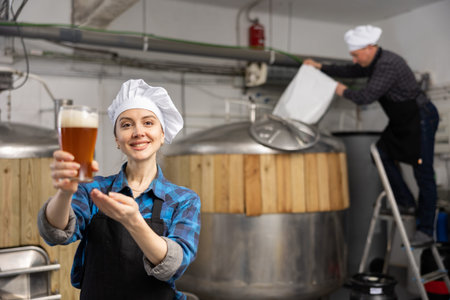 Portrait of female brewer who is standing with glass of beer on her workplace in brew-house
